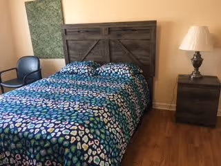 Small bedroom with a double bed covered in a blue-green patterned comforter, a wooden headboard, nightstand with a lamp, a chair, and hardwood floors.