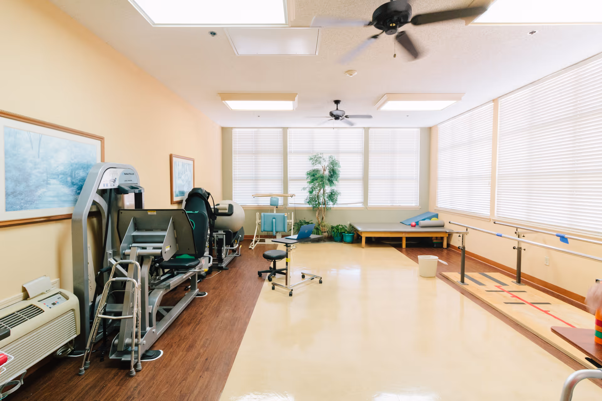 A bright physical therapy or rehabilitation room with exercise equipment, parallel bars for walking practice, a padded therapy table, and a few plants near large windows with blinds. The room has a ceiling fan, framed artwork on the walls, and a walker placed near the exercise machines.