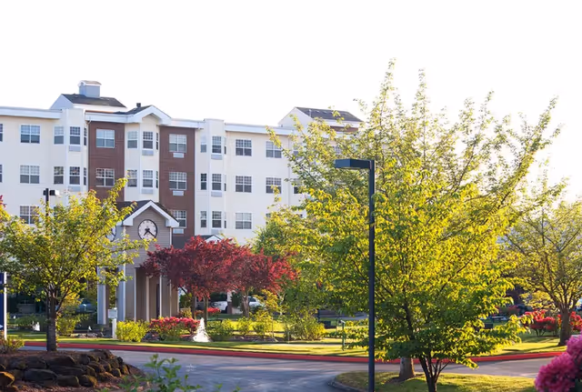 Exterior view of a multi-story senior living facility building with white and brown facade, surrounded by green trees and landscaped garden with a small clock tower structure in front.