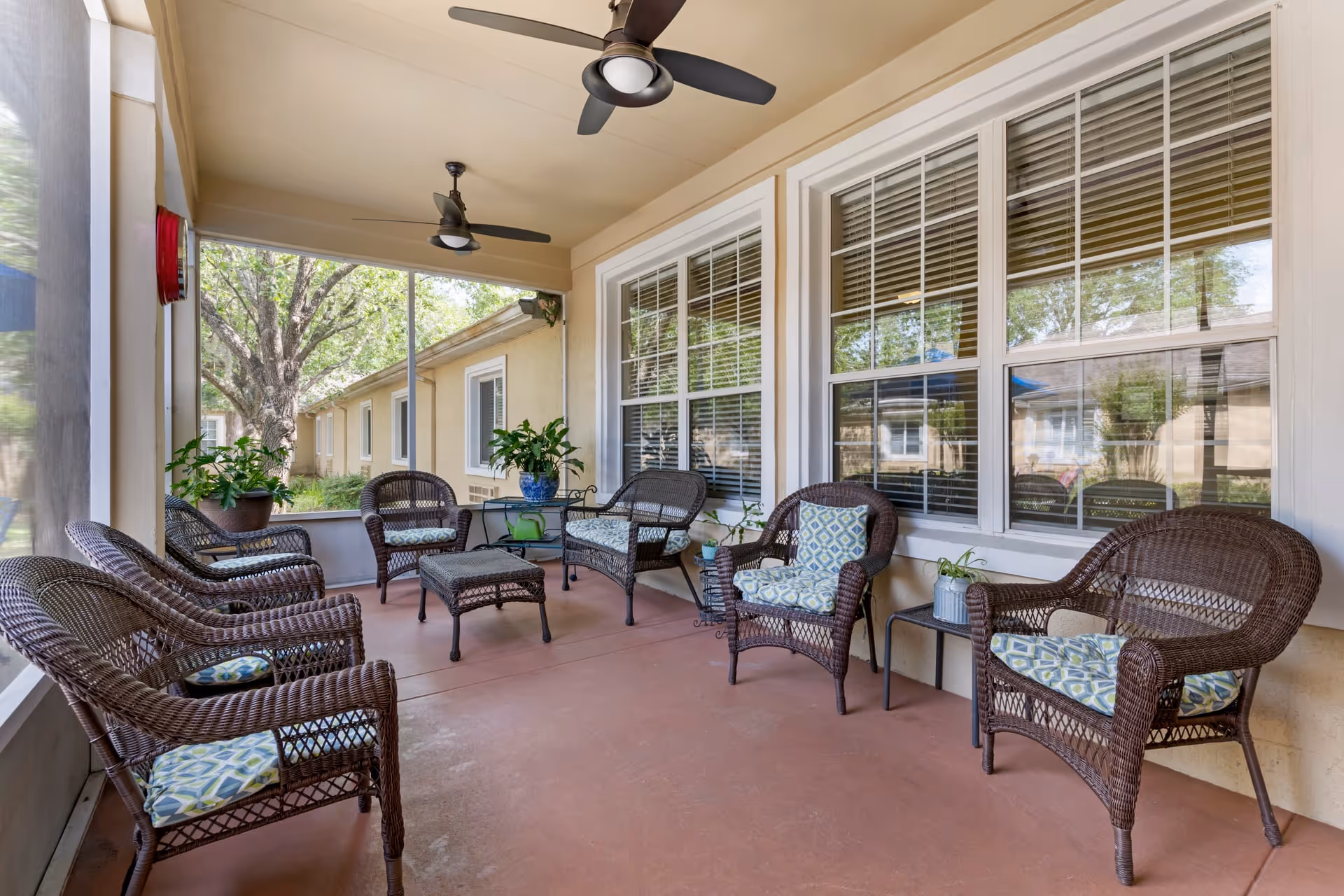 A covered outdoor patio area with several wicker chairs and a wicker loveseat, all with patterned cushions. There are two ceiling fans above, potted plants on small tables, and large windows with blinds on the wall. The patio overlooks a garden area with trees and shrubs.