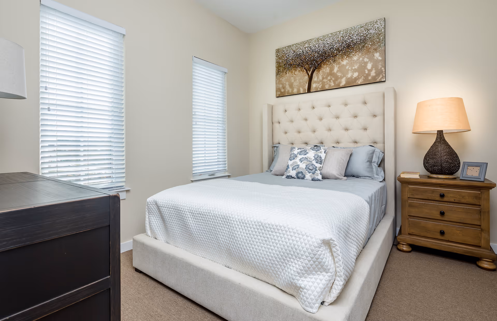 A bedroom with a beige upholstered bed featuring a tufted headboard, white quilted bedspread, and decorative pillows. There are two windows with white blinds on the left wall, a wooden dresser in the foreground, and a wooden nightstand with a lamp and picture frame on the right. A large painting of a tree hangs above the bed.