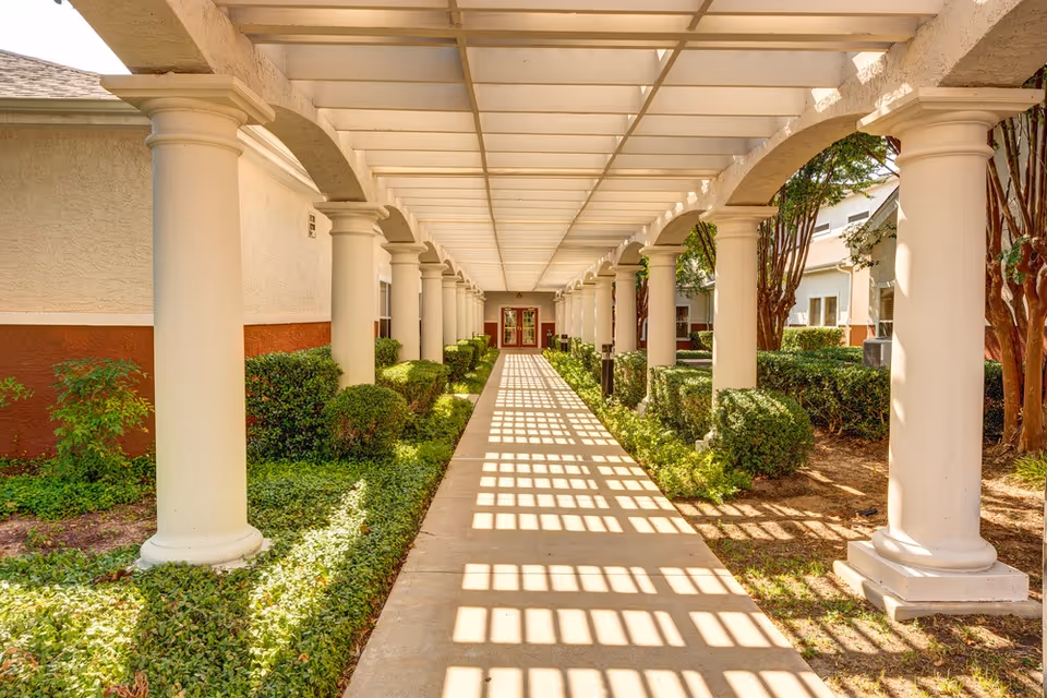 A covered outdoor walkway with white columns on both sides, neatly trimmed bushes and greenery along the path, leading to a building entrance with double doors at the end.