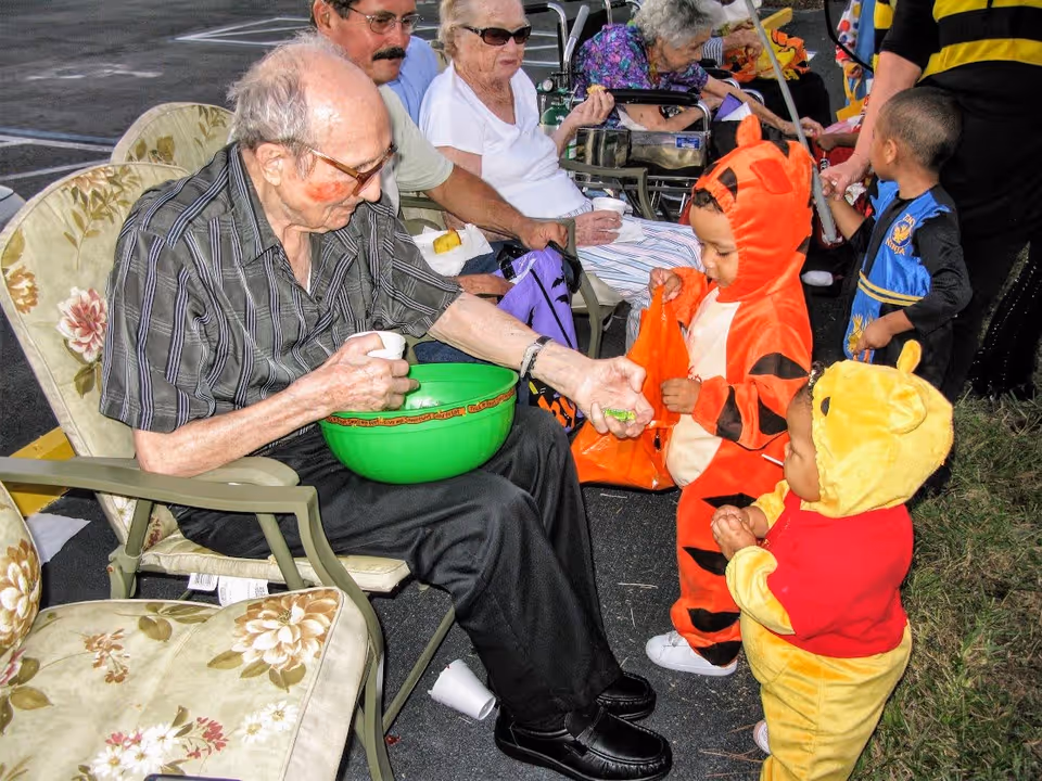 An elderly man sitting in a floral cushioned chair outdoors, handing out candy from a green bowl to two young children dressed in Halloween costumes, one as Tigger and the other as Winnie the Pooh. Other elderly people are seated nearby, and a few more children and adults are visible in the background.