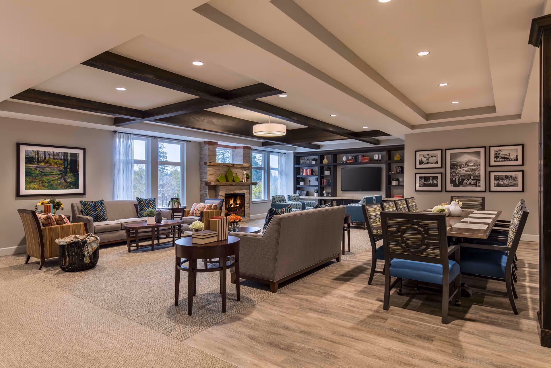 A spacious and well-lit living room area with multiple seating arrangements including sofas and armchairs around a coffee table. There is a fireplace with decorative items on the mantel, large windows letting in natural light, and a wall-mounted television with built-in shelving. To the right, there is a dining table with chairs, artwork on the walls, and a ceiling with exposed wooden beams and recessed lighting.