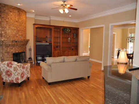 A cozy living room with hardwood floors, a beige sofa facing a wooden entertainment center with a TV, a floral armchair, a brick fireplace on the left, and a ceiling fan with lights above. The room opens into adjacent spaces with beige walls and white trim.