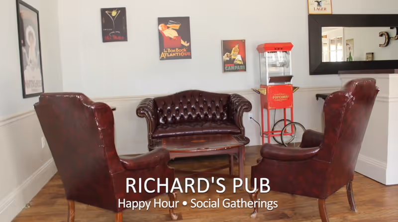 A cozy seating area with three dark brown leather chairs and a matching loveseat arranged around a wooden coffee table. The walls are decorated with vintage-style posters, and there is a red popcorn machine in the corner. A large mirror hangs on the wall to the right.