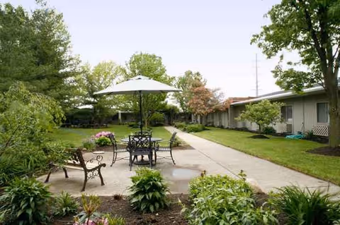 Outdoor courtyard with table and umbrella, walkway, and landscaped garden beside a single-story facility.