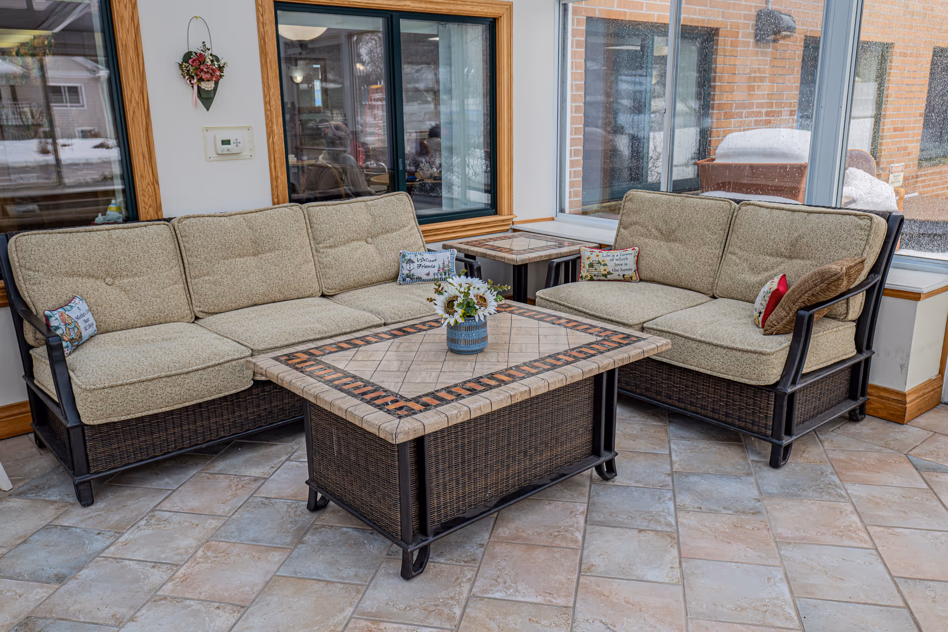 A cozy indoor seating area with two cushioned wicker sofas arranged around a square tiled coffee table. The room has large windows showing an outdoor snowy scene and brick walls outside. Decorative pillows and a small flower arrangement on the table add a homey touch.