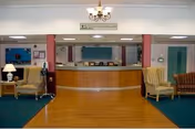 Interior view of a nursing and rehab center reception area with a wooden floor in the foreground and green carpet on the sides. The reception desk is centered with chairs behind it. There are two armchairs and a sofa on the left and right sides of the room. The walls are light-colored with red pillars and a chandelier hanging from the ceiling.