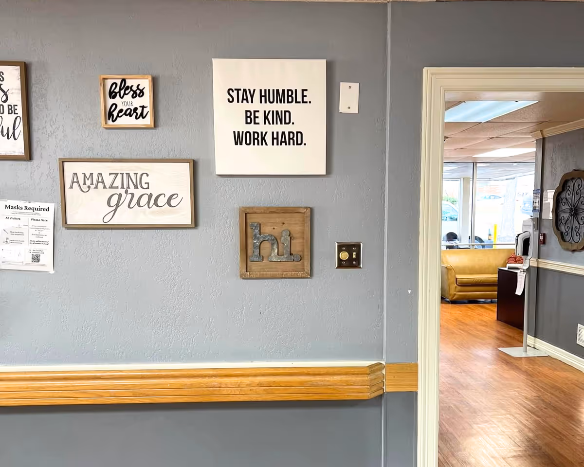 A gray hallway wall decorated with framed inspirational signs and a wooden handrail, with a doorway revealing a seating area.
