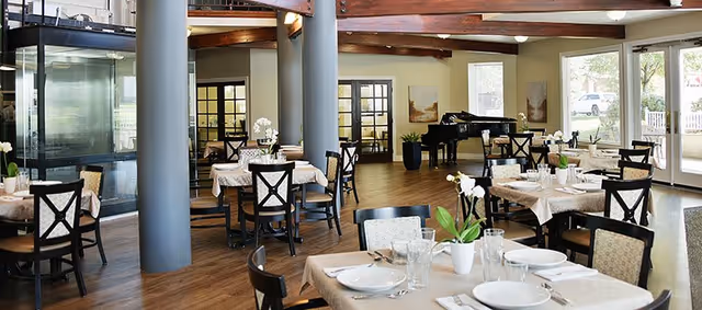 A spacious dining room with multiple tables covered in beige tablecloths, set with white plates, glasses, and cutlery. The room features wooden floors, large windows letting in natural light, a black grand piano in the corner, and modern columns. There are chairs with dark wooden frames and light-colored cushions around the tables.