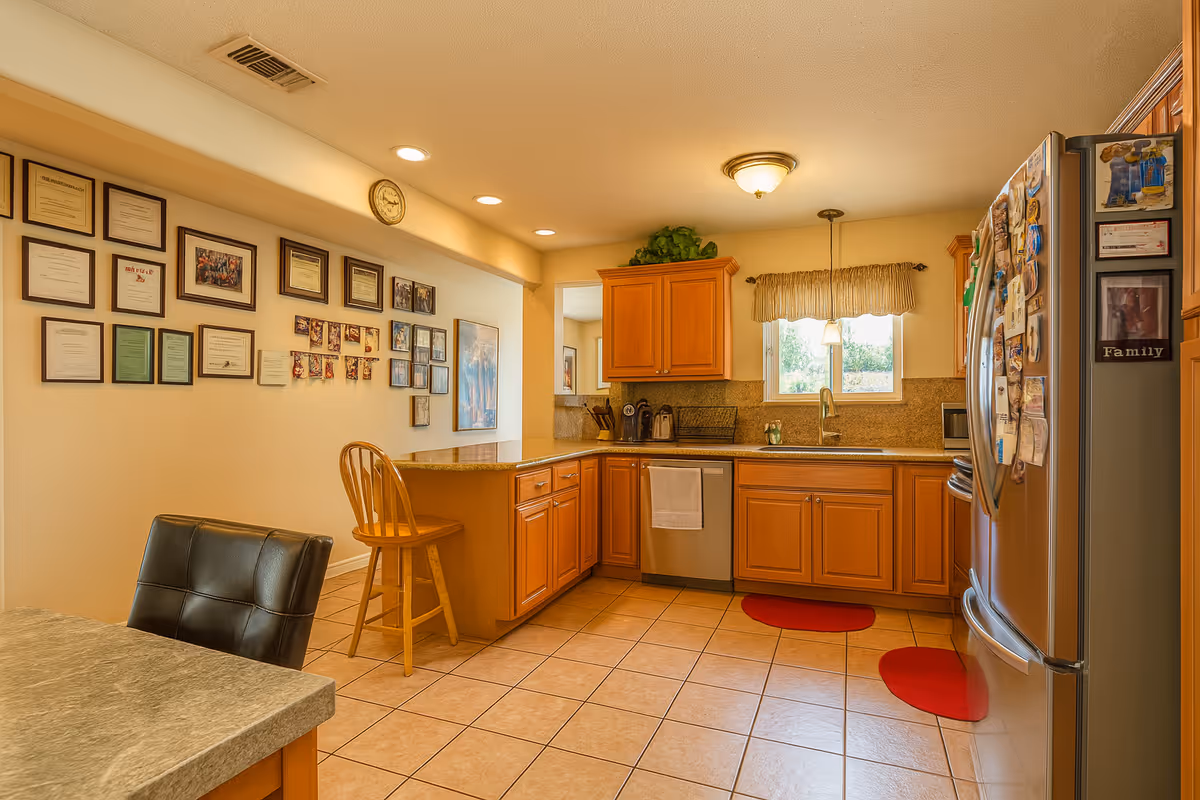 A warm and inviting kitchen with wooden cabinets, a stainless steel refrigerator covered with magnets and photos, a dishwasher, and a sink under a window with a valance. The floor is tiled, and there are two red circular rugs on the floor. On the left side, there is a counter with a wooden chair and a wall decorated with numerous framed certificates and photos. The kitchen is well-lit with recessed lighting and a ceiling light fixture.
