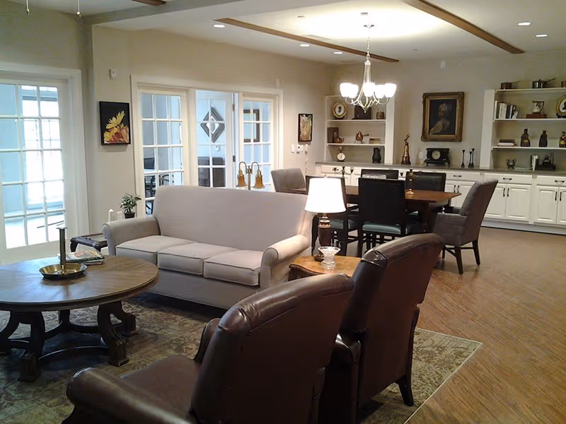A cozy living room area in a senior living facility with a beige sofa, two brown leather armchairs, a round wooden coffee table, and a side table with a lamp. In the background, there is a dining table with chairs, built-in shelves with decorative items, and a chandelier hanging from the ceiling. The room has wooden flooring and light-colored walls with framed artwork.