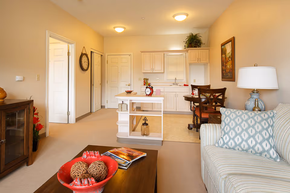 Open-plan living room with a striped sofa, coffee table holding a decorative bowl and magazines, and a small kitchenette with an island in the background.