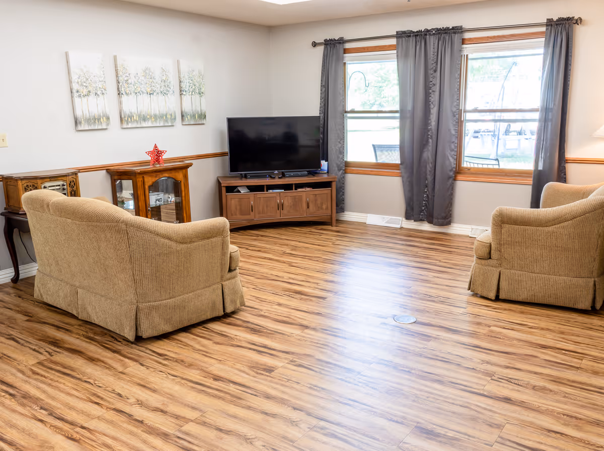 Bright communal living room with two beige armchairs, a TV on a wooden stand, hardwood floors and large windows with gray curtains.