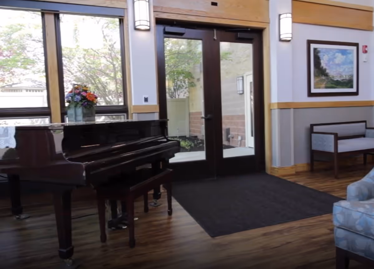 Interior view of a common area featuring a grand piano with a vase of colorful flowers on top, large windows letting in natural light, double glass doors leading outside, wooden flooring, a cushioned bench, and a framed landscape painting on the wall.