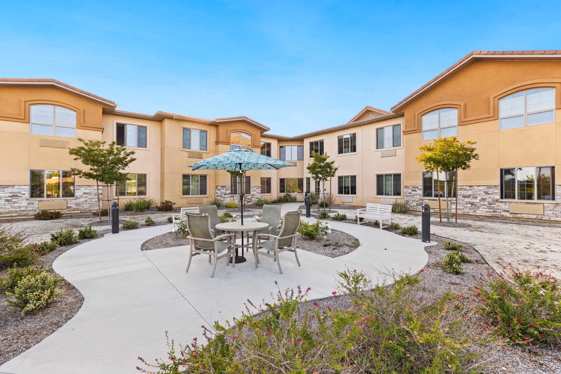 A view of the courtyard at Ivy Park at Murrieta featuring a round table with chairs under a patterned umbrella, surrounded by landscaped gardens and the building's exterior.