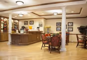 A cozy common area in a senior living facility featuring a wooden counter with a coffee and beverage station, a popcorn machine, and several tables with chairs arranged for socializing or dining. The room has warm lighting, framed pictures on the walls, and decorative plants.