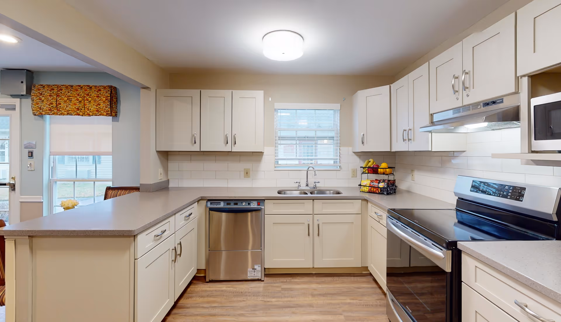 A modern kitchen with white cabinets, a stainless steel dishwasher, an electric stove with a range hood, a microwave, and a double sink under a window. The kitchen has light-colored countertops and wood flooring. There is a fruit basket on the counter and a window with blinds above the sink. The kitchen opens to an adjacent room with a window and a door.