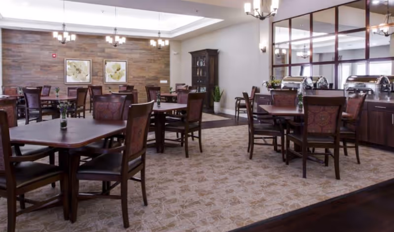 A dining room in an assisted living facility with several wooden tables and chairs arranged neatly. The room features a patterned carpet, a wooden accent wall with two framed floral artworks, and a cabinet with glass doors. There are buffet warmers on a sideboard along one wall, and modern light fixtures hang from the ceiling.