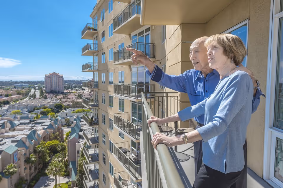 An elderly couple stands on a balcony of a high-rise building, with the man pointing out towards the cityscape and the woman holding the railing. The background shows a clear blue sky and a view of nearby buildings and streets.