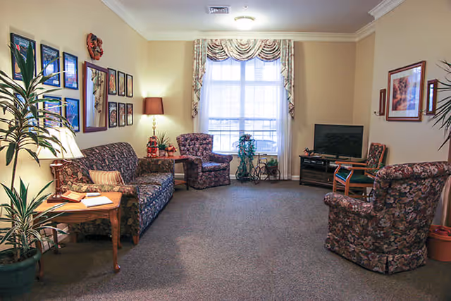 A cozy living room with floral patterned sofa and armchairs arranged around a window with draped curtains. The room features framed pictures on the walls, a television on a stand, a side table with a lamp, and several potted plants.