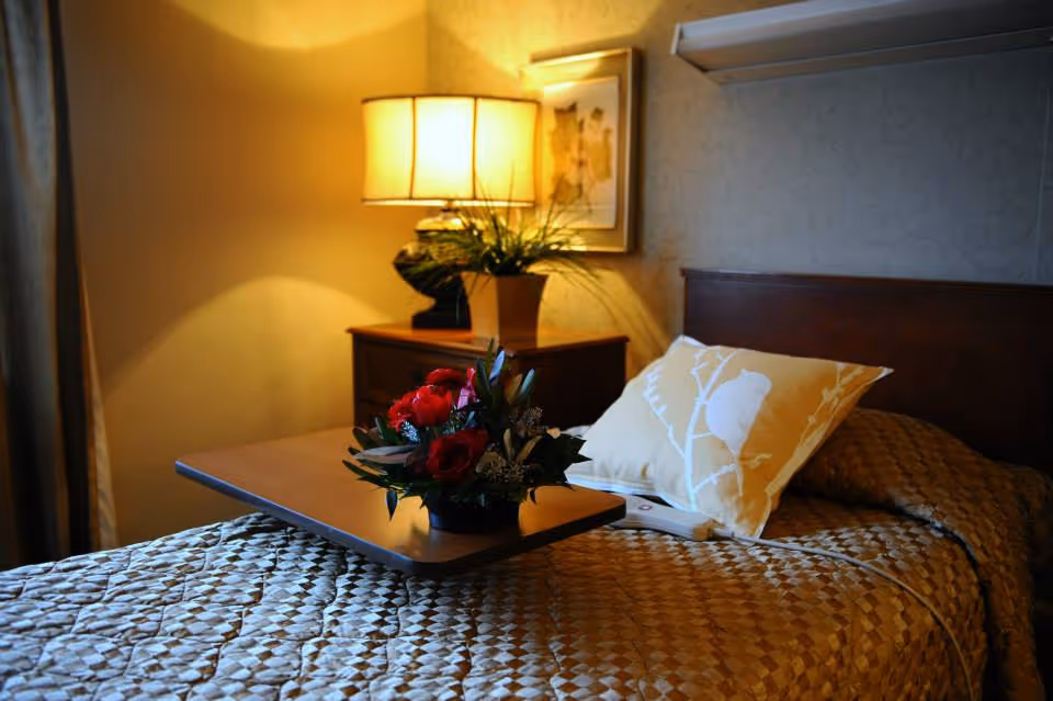 A cozy bedside scene with a made single bed, decorative pillow, bedside table lamp and a tray holding a bouquet of red flowers.