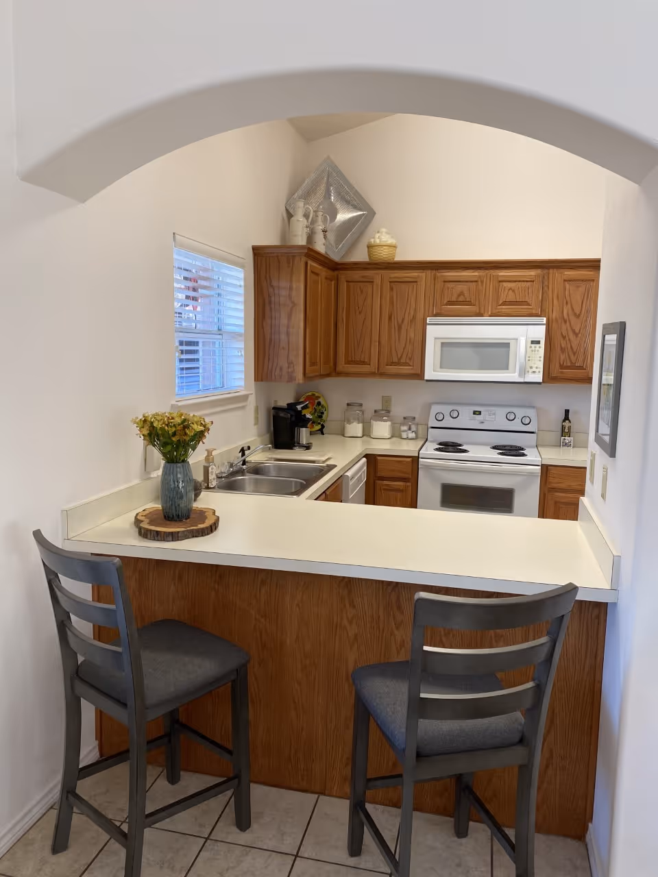 Small kitchen area with wooden cabinets, a white countertop, and white appliances including a microwave and stove. There is a double sink with a coffee maker nearby, and two gray cushioned bar stools at the counter. A window with blinds is on the left wall, and decorative items are placed on top of the cabinets and countertop.
