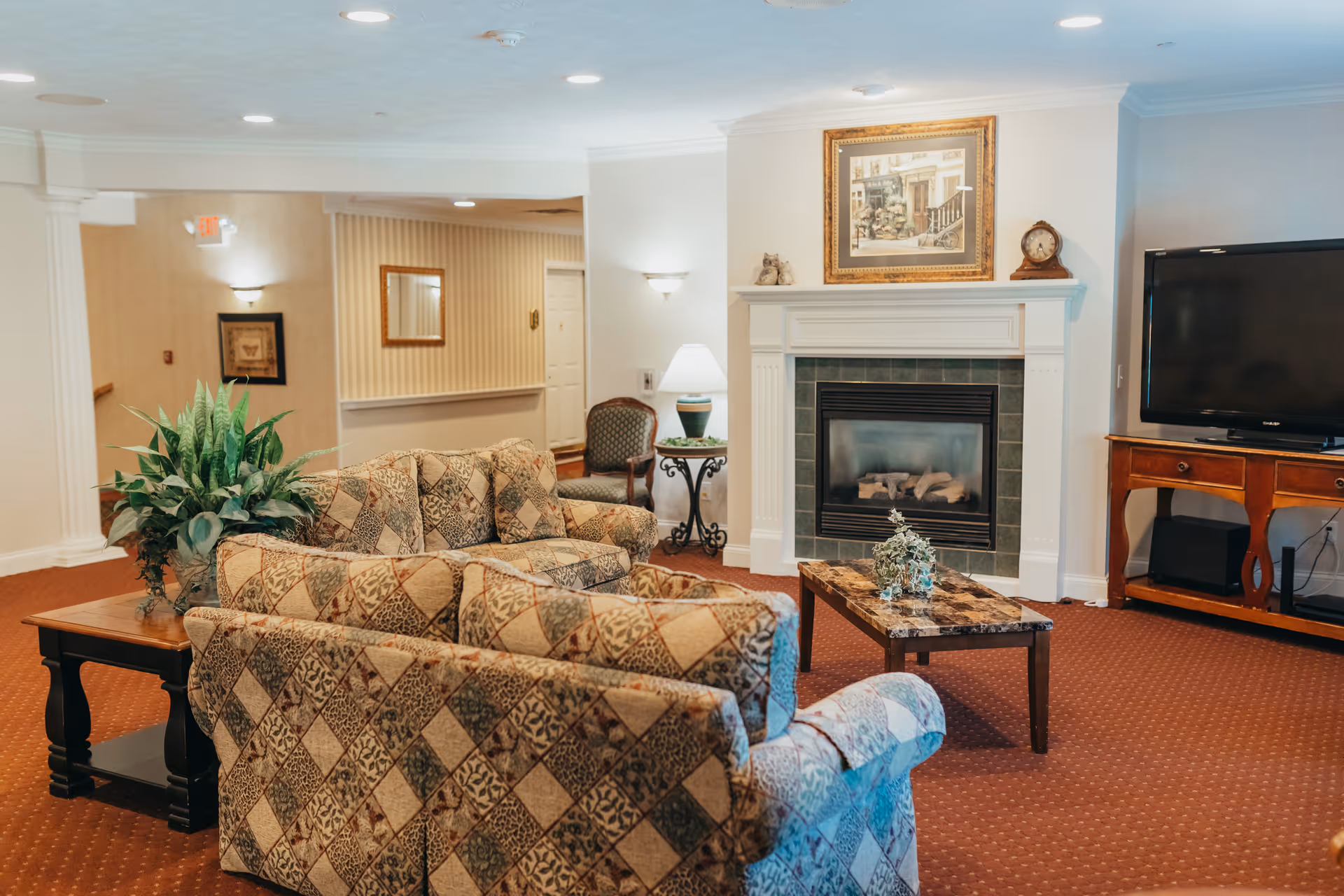 A cozy living room area in a senior living facility featuring patterned sofas, a coffee table with a small plant, a fireplace with a framed picture above it, a side table with a lamp, and a flat-screen TV on a wooden stand. The room has warm lighting and carpeted floors.