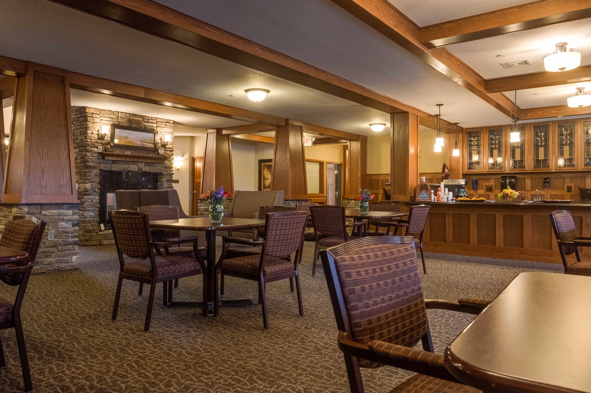 A cozy dining area in a senior living facility with multiple tables and chairs arranged on a carpeted floor. The room features wooden beams on the ceiling, a stone fireplace with a painting above it, and a counter area with cabinets and pendant lights. Vases with flowers are placed on the tables, creating a warm and inviting atmosphere.