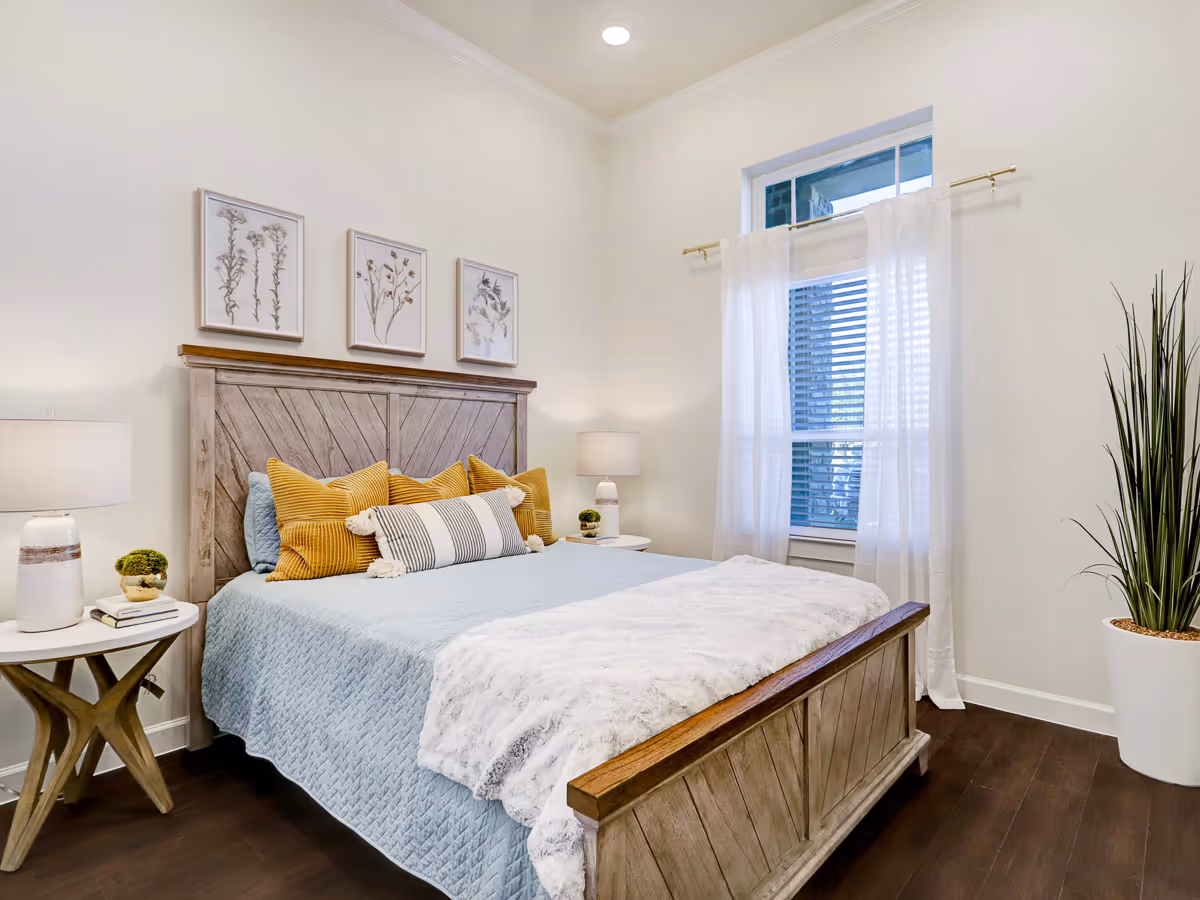 Well-lit bedroom featuring a wooden bed with blue bedding and yellow pillows, bedside tables with lamps, framed botanical prints, and a window with sheer curtains.