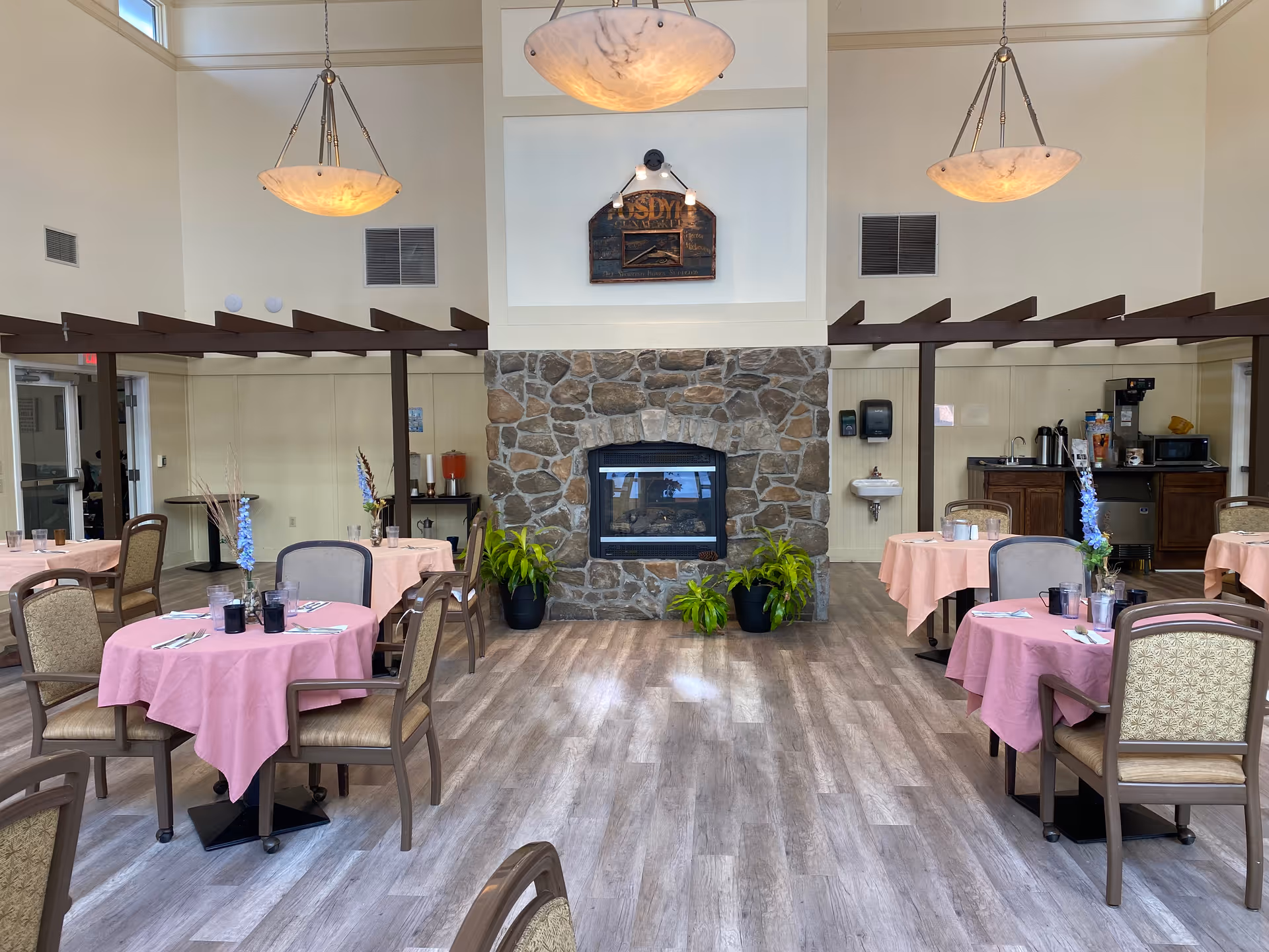 A dining room in an assisted living facility with round tables covered in pink tablecloths, each set with glasses, utensils, and a small flower arrangement. The room features a stone fireplace in the center with green plants at its base, wooden beams on the ceiling, and three hanging pendant lights. In the background, there is a small kitchenette area with a coffee machine, microwave, and sink.