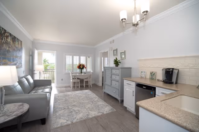 A bright and airy living space featuring a gray leather sofa on the left, a light-colored area rug on the wooden floor, and a small dining table with four chairs near a window with white shutters. On the right side, there is a kitchen counter with a coffee maker, a dishwasher, and a sink. The walls are painted light gray and decorated with framed pictures and a large landscape painting. A chandelier hangs from the ceiling.