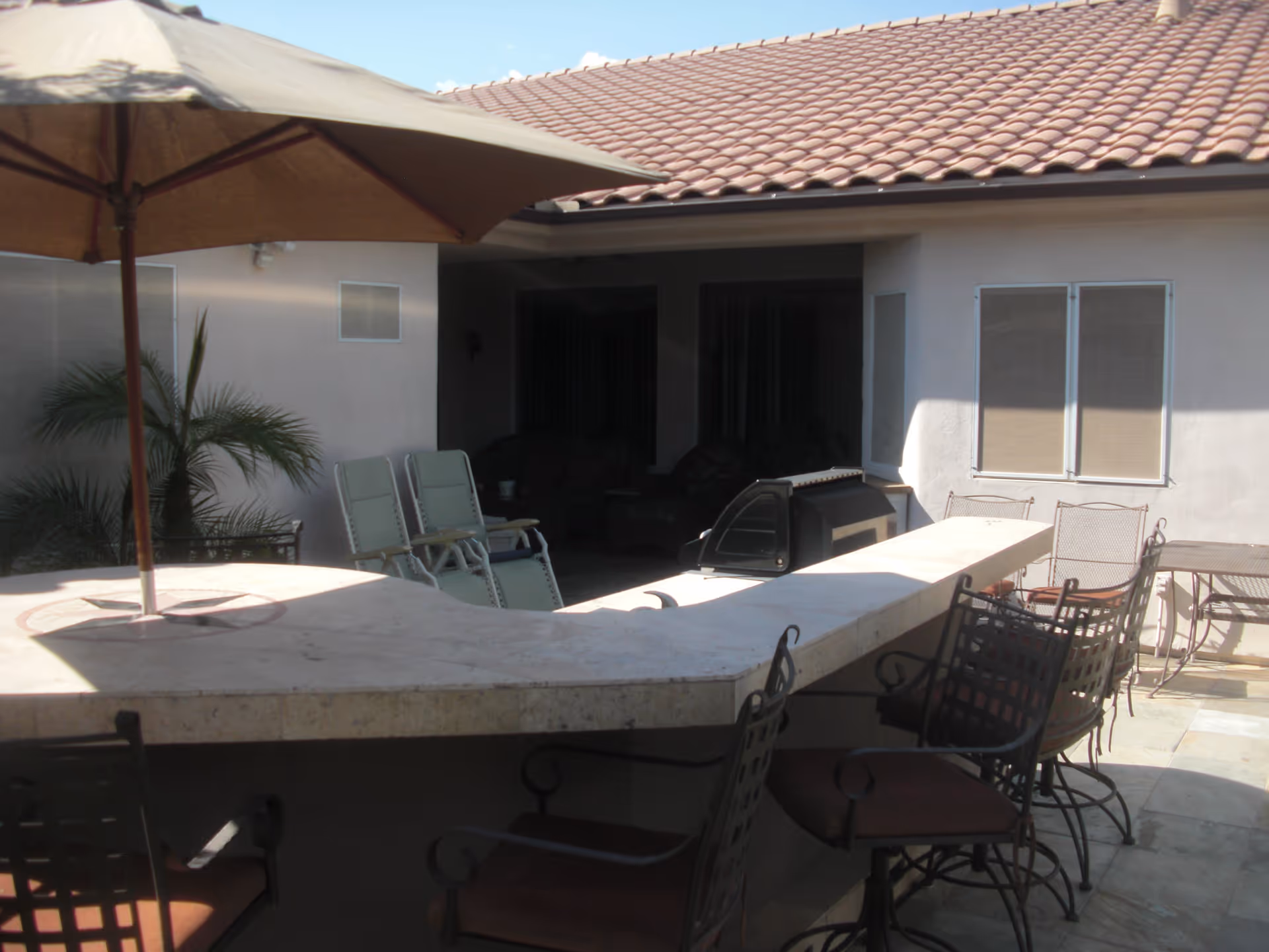 Outdoor patio area with a curved stone countertop bar, several metal chairs with cushions, a large beige umbrella providing shade, and a barbecue grill. The patio is adjacent to a building with a tiled roof and windows with closed blinds. There are also some lounge chairs and a small palm plant visible.