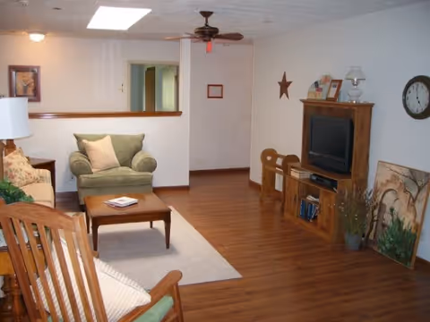 A cozy living room with wooden flooring, a green armchair with a pillow, a wooden rocking chair, a coffee table with a book, a wooden TV stand with a television, books, and decorative items. The walls are light-colored with a star decoration, a clock, and framed pictures. A ceiling fan with a light is mounted on the ceiling.