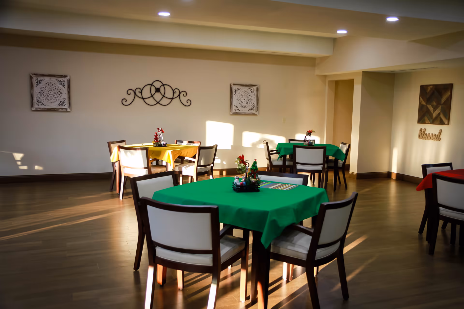A dining room with several square tables covered with colorful tablecloths in green, yellow, and red. Each table is surrounded by four chairs with white cushions and dark wooden frames. The room has wooden flooring, light-colored walls decorated with framed art and a metal wall ornament, and ceiling lights. Small holiday-themed centerpieces are placed on each table.