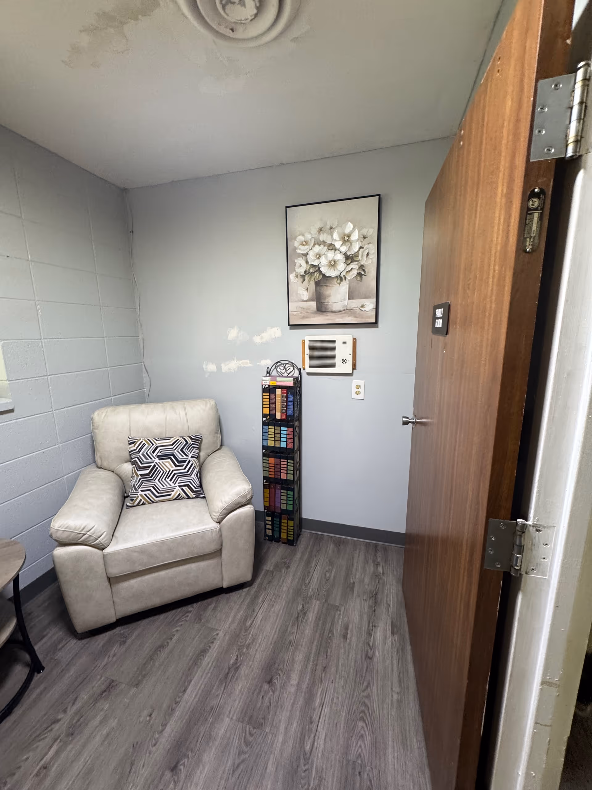 Small sitting room with a beige recliner and patterned pillow, a side table, tall book rack, and floral wall art.