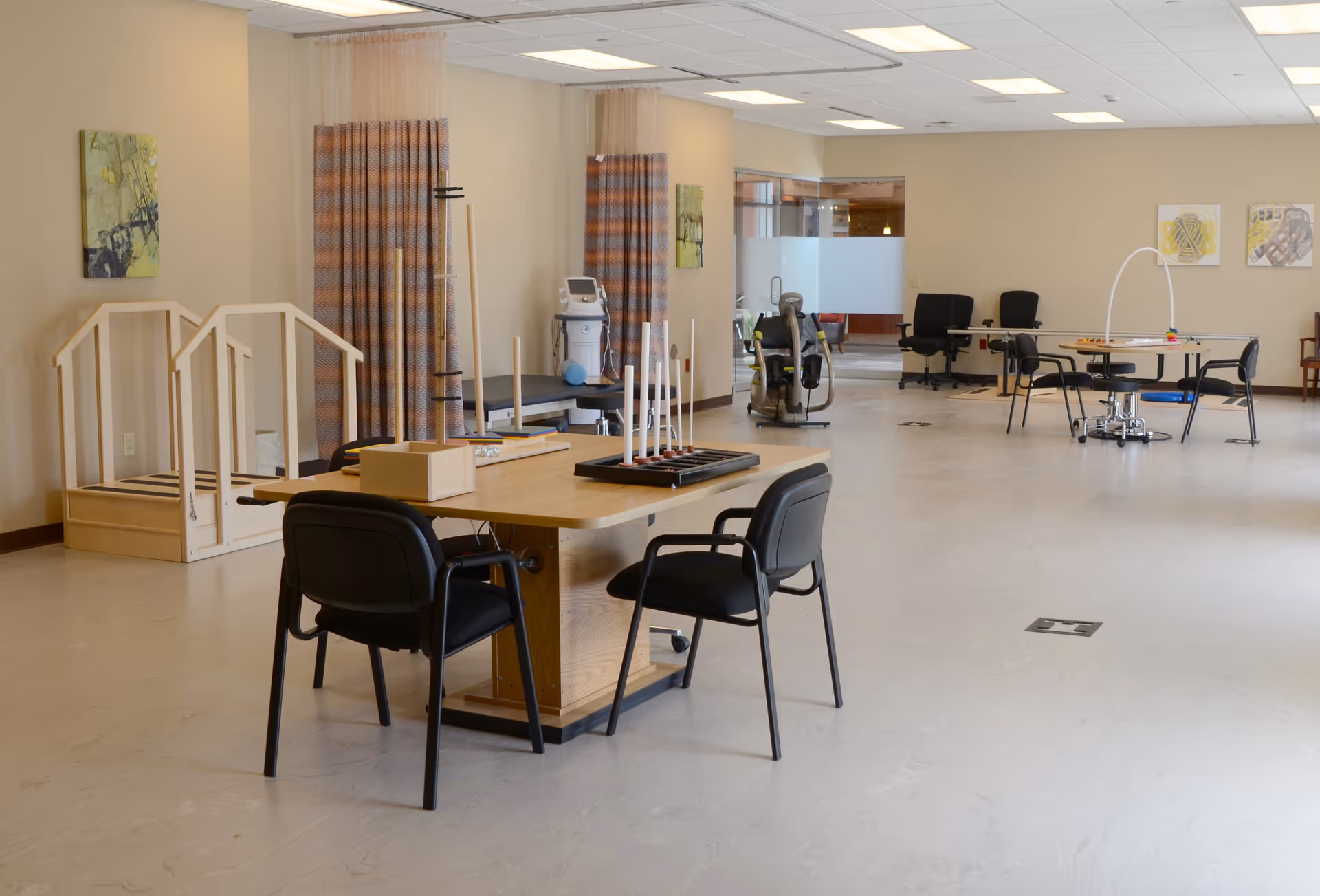 A spacious senior activity and therapy room with tables and chairs, exercise and rehabilitation equipment, and wooden practice stairs.
