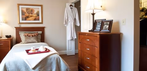 A neatly made single bed in a senior living bedroom with a tray on the bed, a wooden dresser with a lamp and framed photos, and a robe hanging on the door.