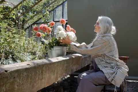 An elderly woman seated outdoors arranging potted flowers on a stone ledge.