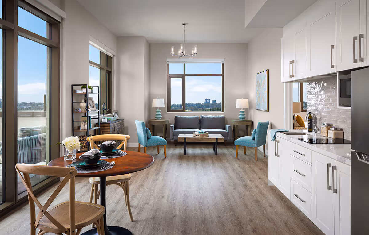 Open-plan living area with a kitchen on the right, a round dining table in the foreground, and a sofa with chairs facing large windows and a balcony.