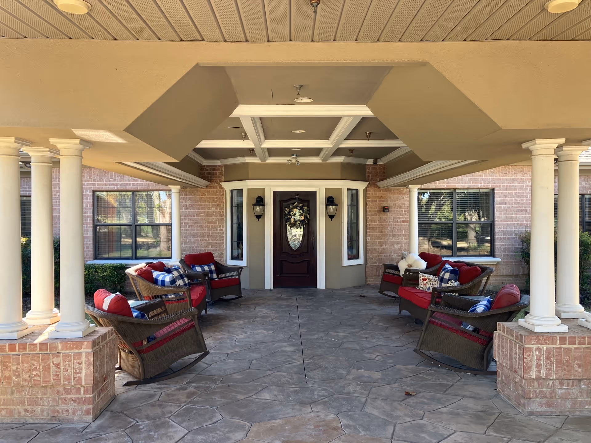 Covered front entrance patio with wicker chairs and red cushions arranged around a walkway leading to a decorative front door.