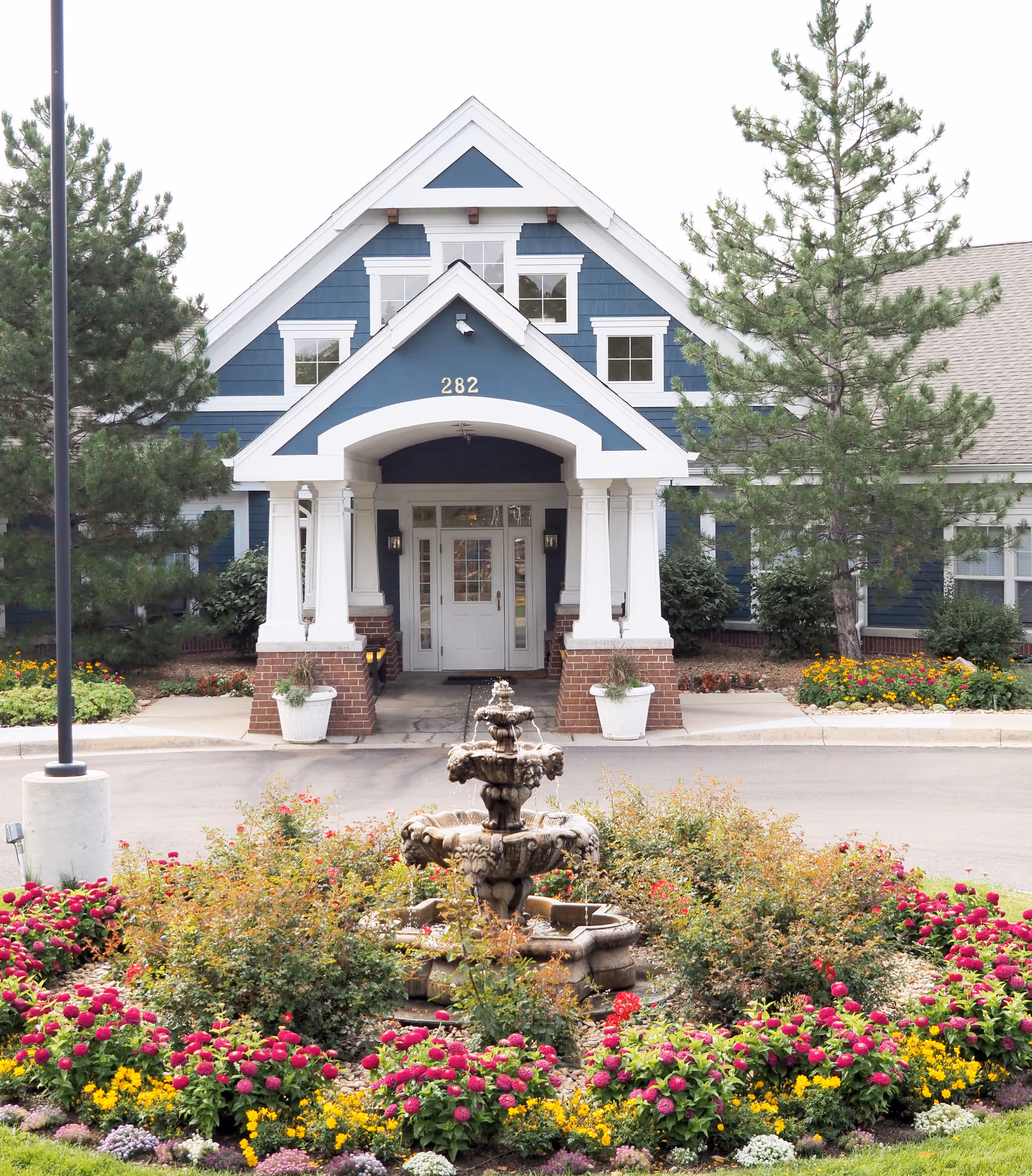 Front entrance of a blue and white building with the number 282 above the door, surrounded by trees and colorful flower beds with a multi-tiered stone fountain in the center.