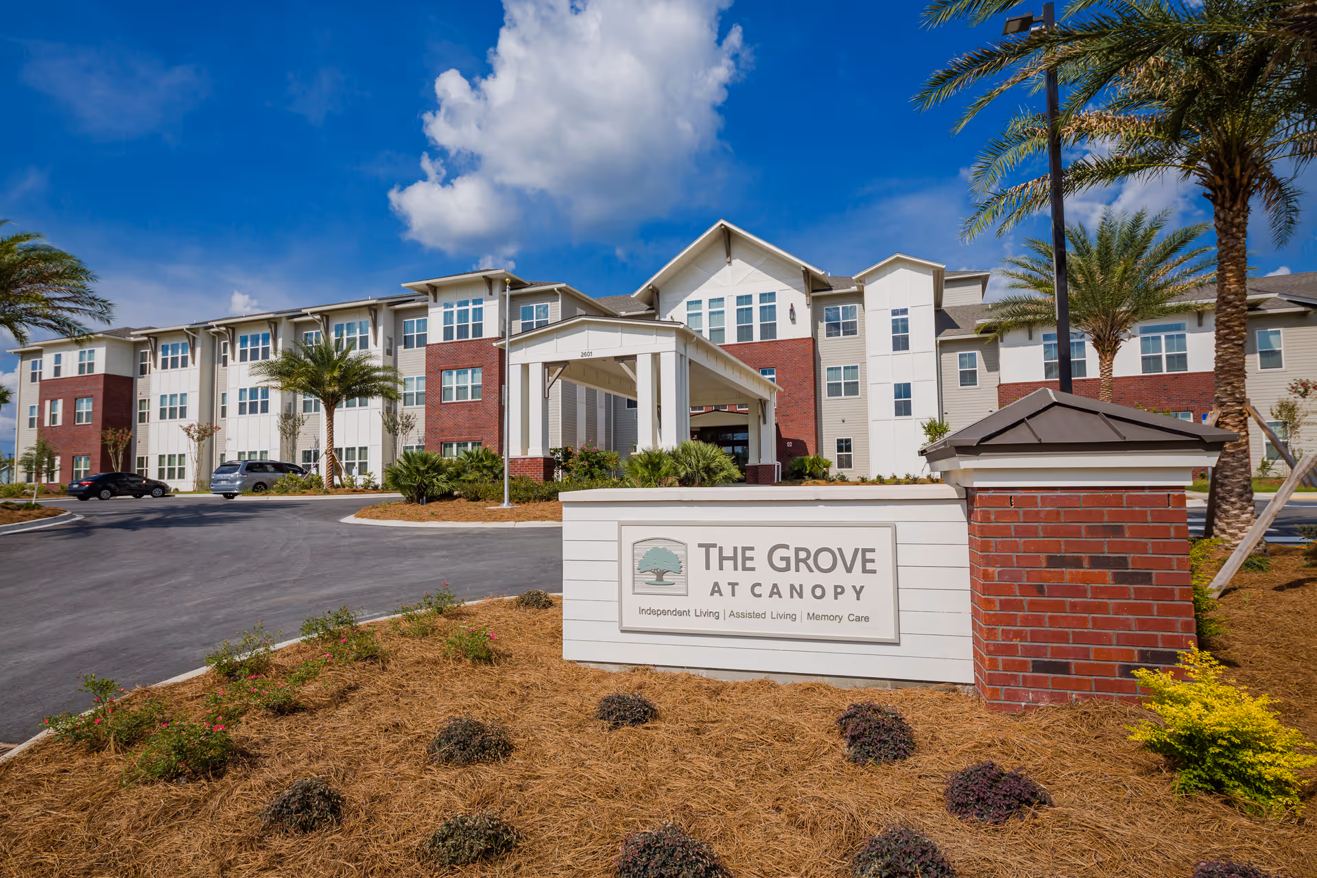 Exterior view of The Grove at Canopy senior living facility showing a large three-story building with a covered entrance, palm trees, landscaped grounds, and a sign in the foreground displaying the facility name and services offered.