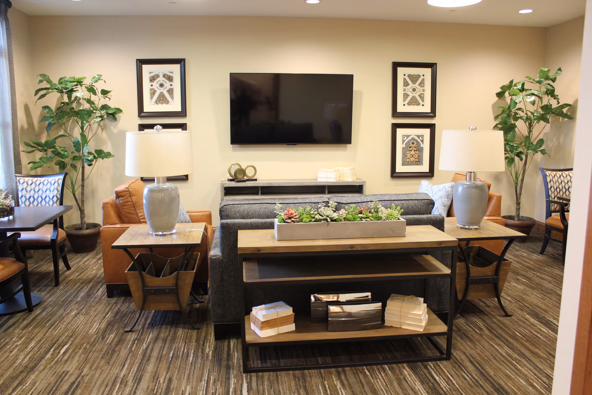 A cozy living room area with a gray sofa facing a wall-mounted flat screen TV. On either side of the sofa are wooden side tables with gray lamps. Behind the sofa is a wooden console table with decorative items including a planter with succulents and stacks of books. The room has patterned carpet flooring, two potted plants in the corners, framed artwork on the wall, and additional seating with chairs and a table visible on the sides.