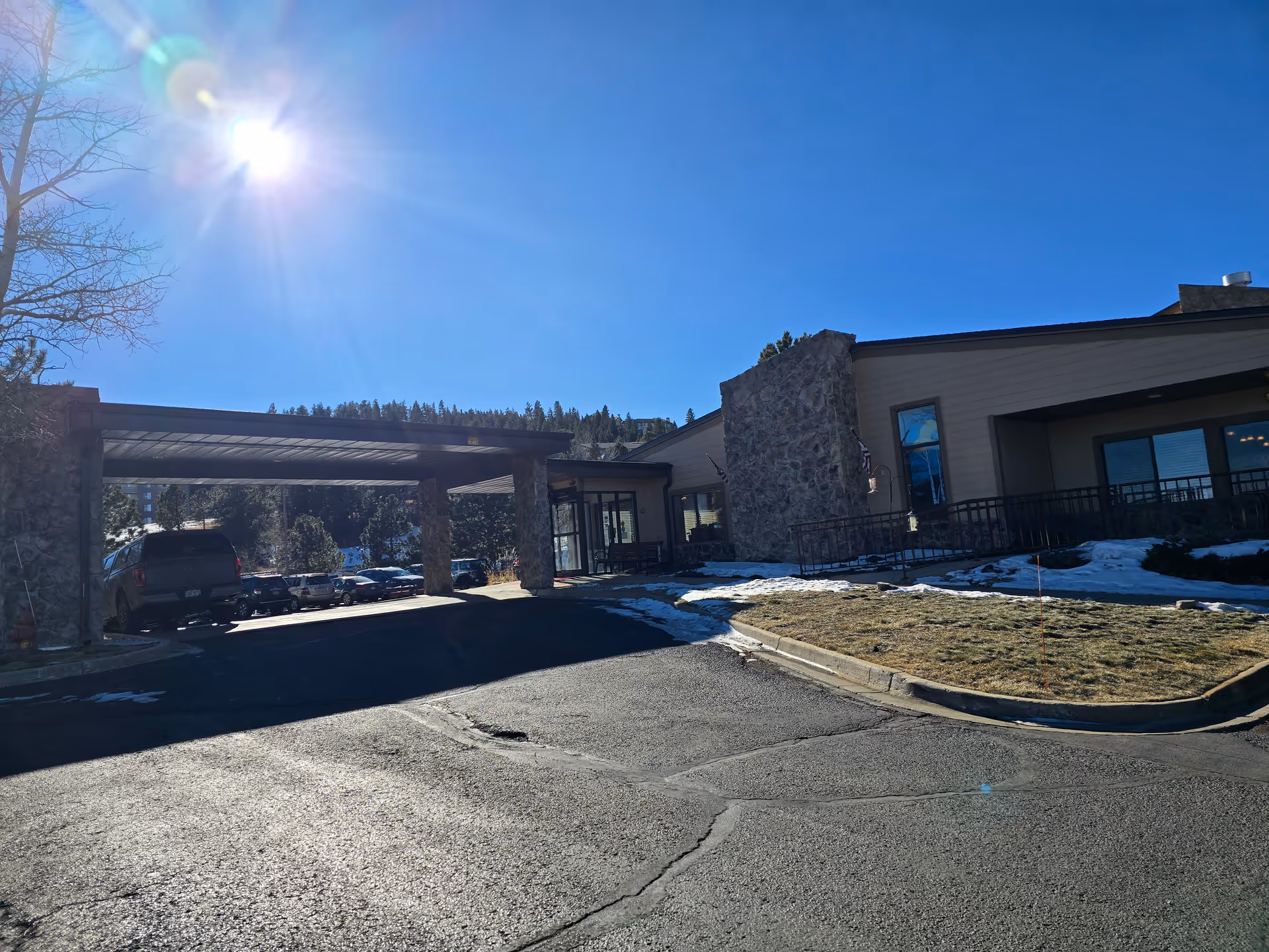 Exterior view of the Life Care Center of Evergreen building on a sunny day with a clear blue sky. The building features stone pillars and a covered entrance area with several parked cars visible. There is some snow on the ground and a grassy area near the entrance.