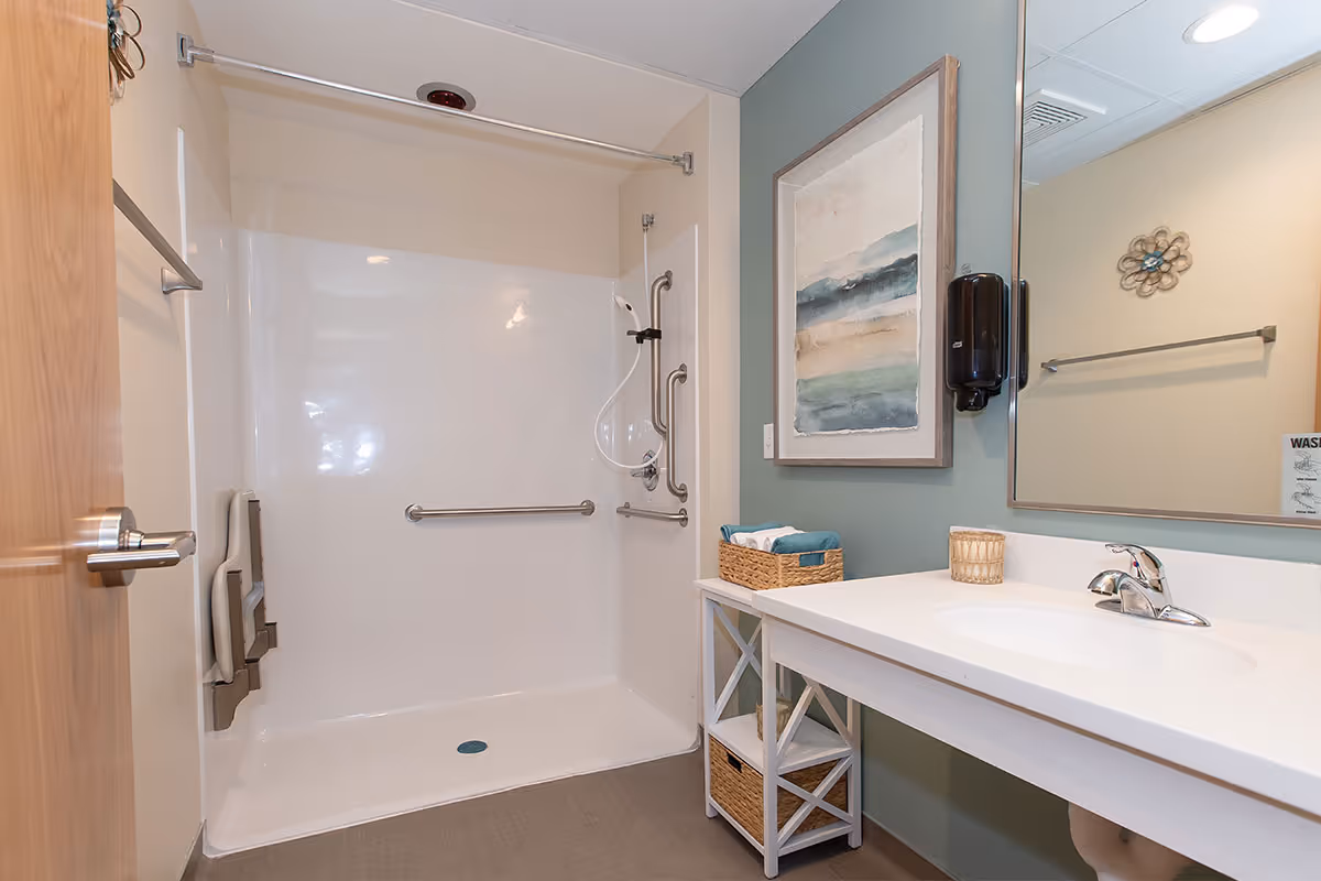 A clean and accessible bathroom with a walk-in shower featuring grab bars and a fold-down seat. There is a white vanity with a sink, a basket with towels, a soap dispenser mounted on the wall, a large mirror, and a framed abstract painting on the wall.