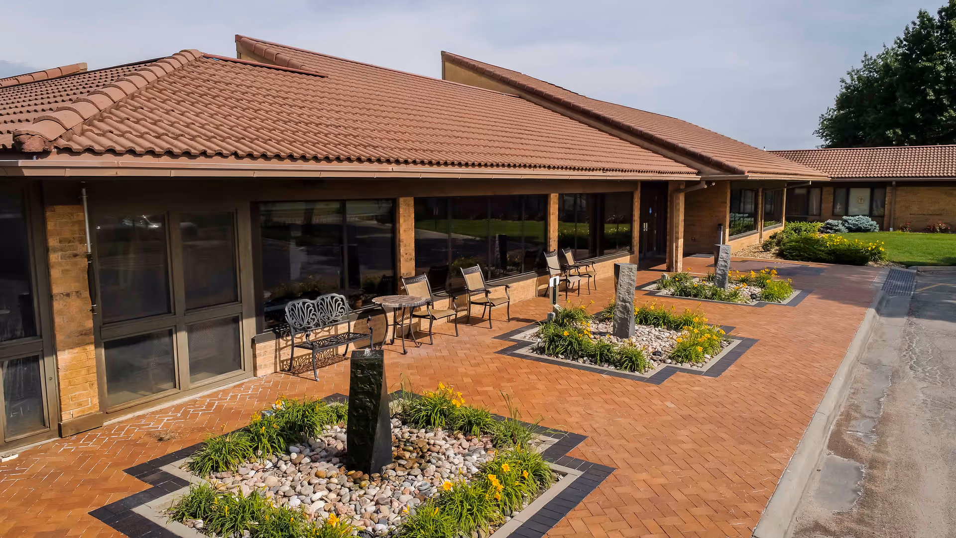 Outdoor patio area of a single-story building with a tiled roof, featuring brick-paved walkways, small landscaped garden beds with rocks and plants, several metal chairs and tables, and large windows along the building's exterior.