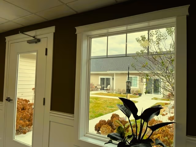View from inside a room looking out through a large window and a glass door onto a courtyard with a pathway, plants, and a building with multiple doors and windows.
