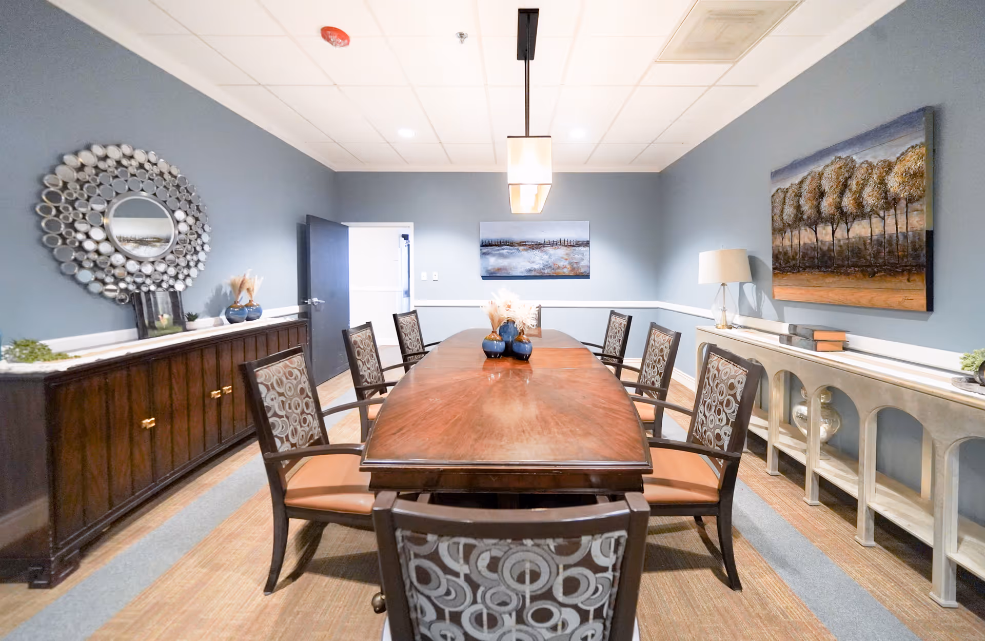 A bright dining room with a long wooden table, upholstered chairs, sideboards and wall art against blue-gray walls.